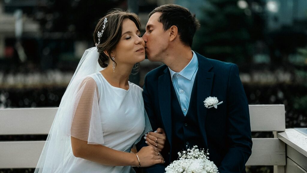 Bride and groom share a tender kiss on a park bench during a wedding day shoot, creating a romantic moment.