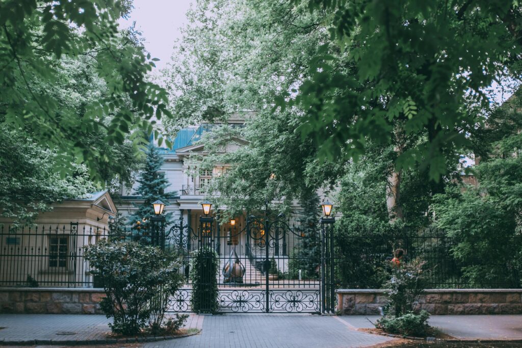 Exterior of stylish classic styled mansion with forged fence and elegant street lamps surrounded by lush green trees in daytime